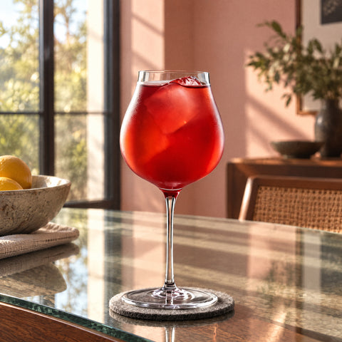 Cocktail rouge, gros glaçon et fruit sombre dans le verre à vin Cheminée sur sous-verre en feutre gris, mur rose poudré