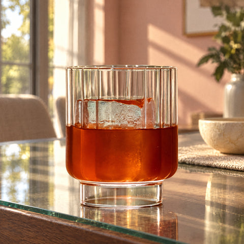 Cocktail ambré et gros glaçon dans le verre à whisky Ionique, mur rose poudré et hautes fenêtres en fond flou