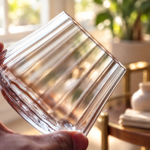 Gros plan sur la paroi côtelée et le buvant du verre à whisky Corset tenu du bout des doigts, desserte en laiton floue