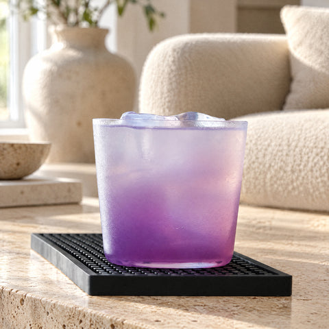 Verre à whisky Plume embué garni d'un cocktail violet et de glaçons, canapé bouclé crème et vase en céramique flous