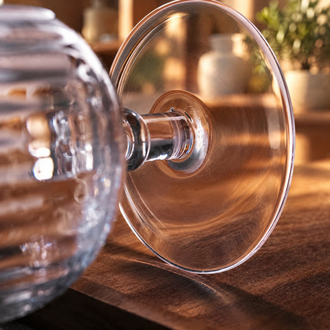 Gros plan sur la tige annelée et la base ronde du verre long drink à pied Arpeggio, reflets ambrés, planche en bois