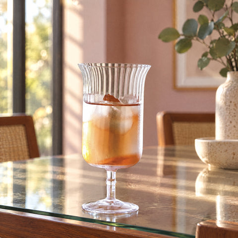 Verre long drink à pied Arpeggio rempli d'une boisson ambrée avec glaçons, mur rose poudré et grandes fenêtres floues