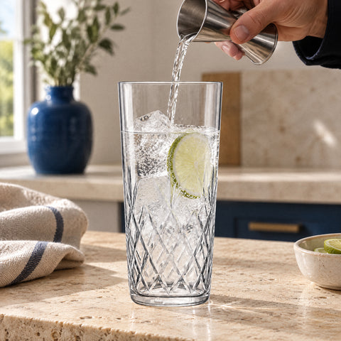 Main versant un gin tonic au jigger dans le verre long drink Palmette sur glaçons et citron vert, comptoir en travertin
