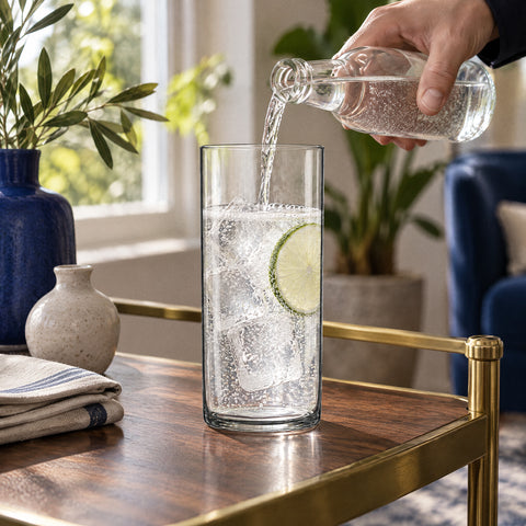 Tonic versé d'une main dans le verre long drink Aplomb sur gin, glaçons et citron vert, desserte en noyer et laiton