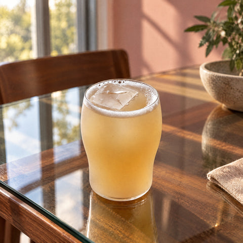 Verre nonic Cambrure vu en plongée, rempli de bière pâle trouble avec glaçon et buée, mur rose poudré et hautes fenêtres